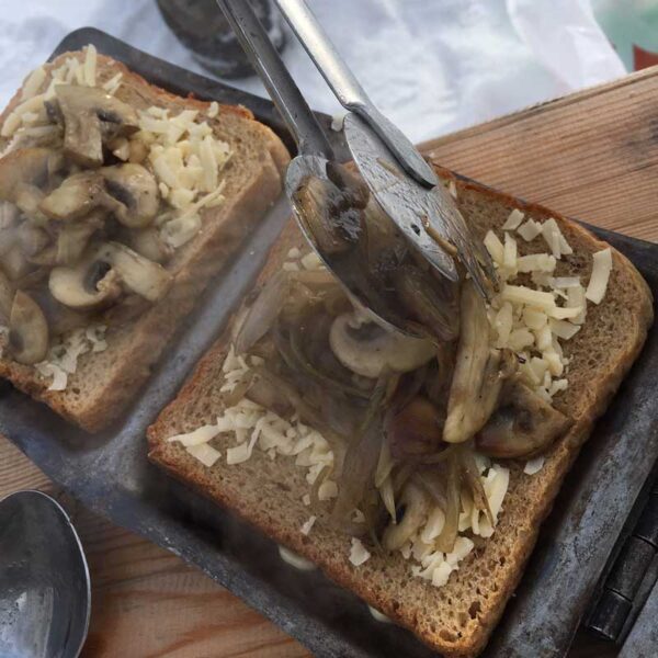 Ett rostfritt serveringsbestick som också kan användas som grilltång används här för att portionera stekt svamp och lök på bröd i ett mackjärn.