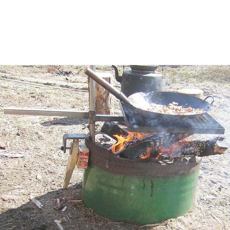 Grillgaller med grillytan 30x40cm är monterat med ett cementfäste på kanten på ett oljefat. Oljefatet står på en sandstrand och i det brinner en eld. På grillgallet står en wokpanna med mat i.