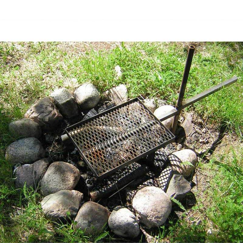 Foto visar en eldstad på marken med stenar i en ring. Ett rektangulärt grillgaller från Eldmark är monterat på ett markrör som är nerslaget i marken mellan stenarna.