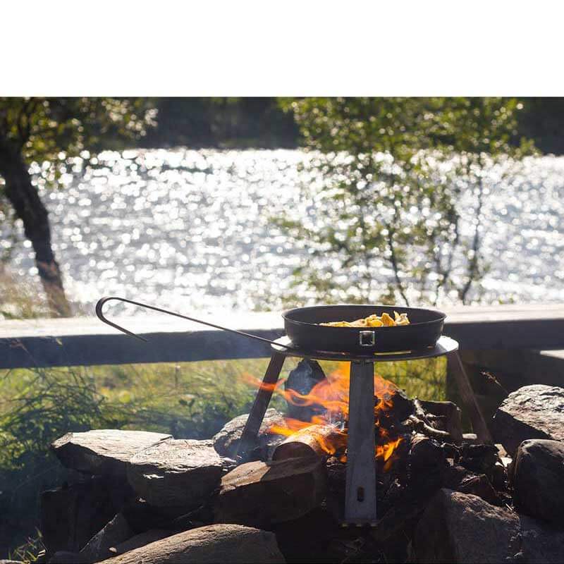 Ett stativ som kallas trefot står över en öppen eld med en sittbänk och en solglittrande sjö o balgrunden. På trefoten finns en stekpanna med mat som puttrar i.