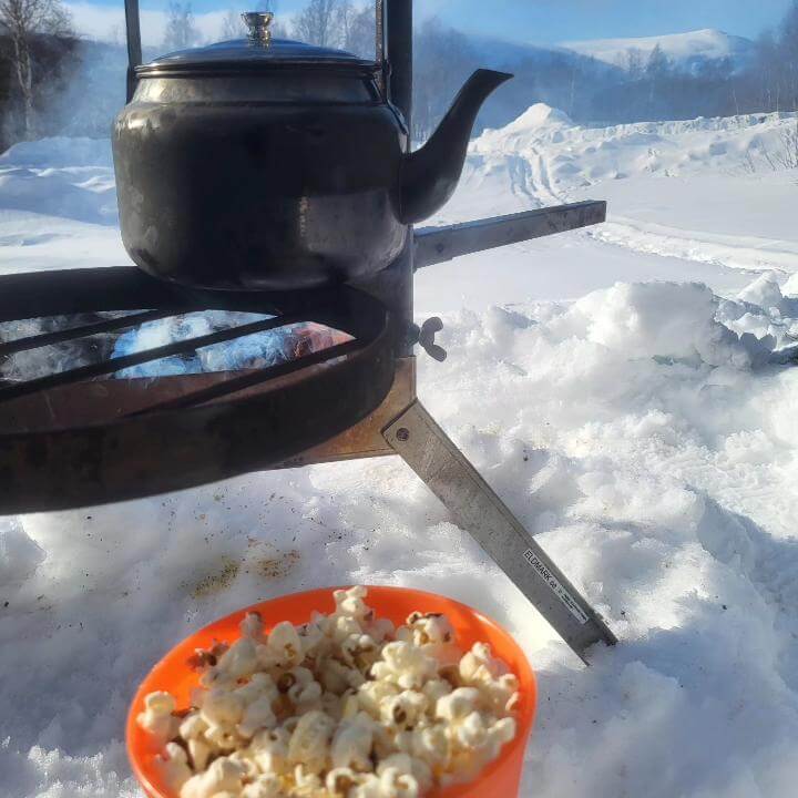 En sotig kaffepanna står på Eldbox och en orange skål med popcorn står under. I bakgrunden ett vinterlandskap.