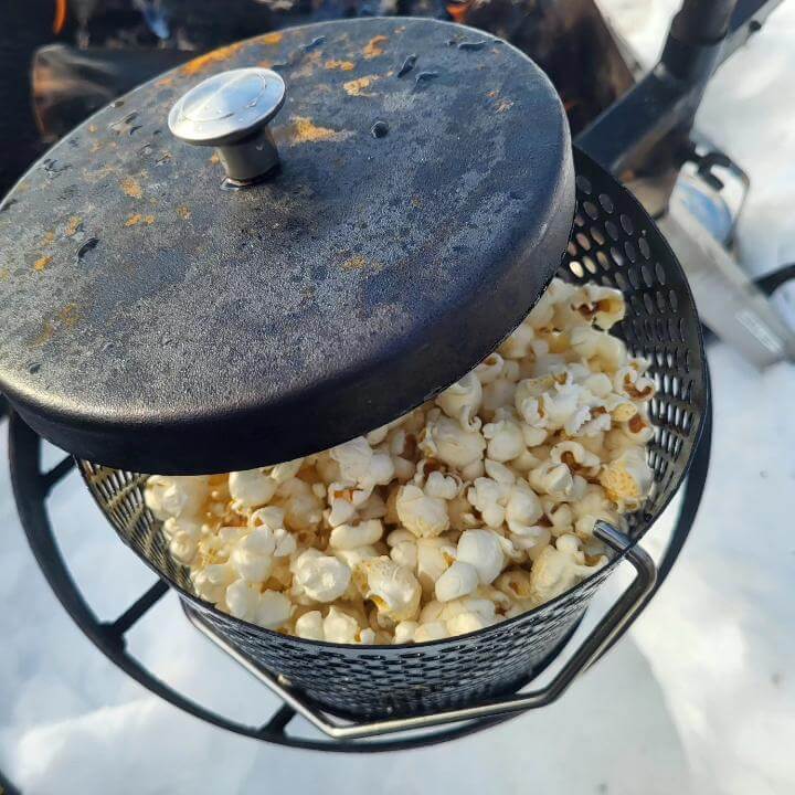 En popcorngryta med halvöppet lock står på eldbox. Popcorn poppade över öppen eld.