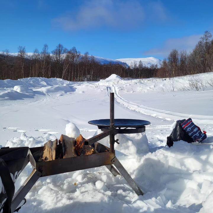 Eldbox med en tänd eld och en stekhäll ligger på hållaren. Vinterlandskap i bakgrunden.