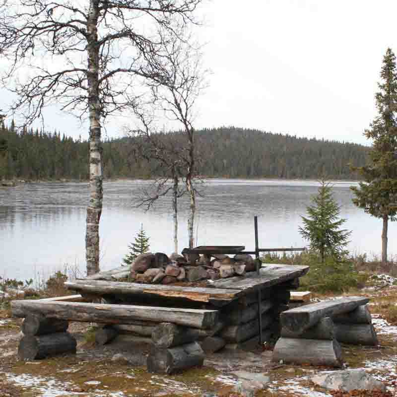 En timrad eldstad och bänkar av stockar runt omkring. Ett höj och sänkbart galler är monterat. I bakgrunden en sjö och ett berg. en björk som tappat löven.