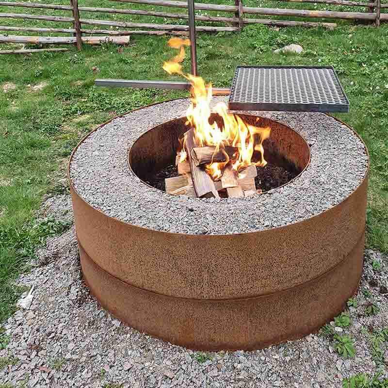 En grillplats av två cortenringar som står på marken. Ringarna har olika diameter och utrymmet mellan dem är fyllt med grus. Ett markrör är nedslaget i grusbädden och på röret är ett grillgaller 50x40 från Eldmark monterat. I centrum brinner en eld.<br />Corten är en sorts rosttrögt stål som blir rostrött till färgen.