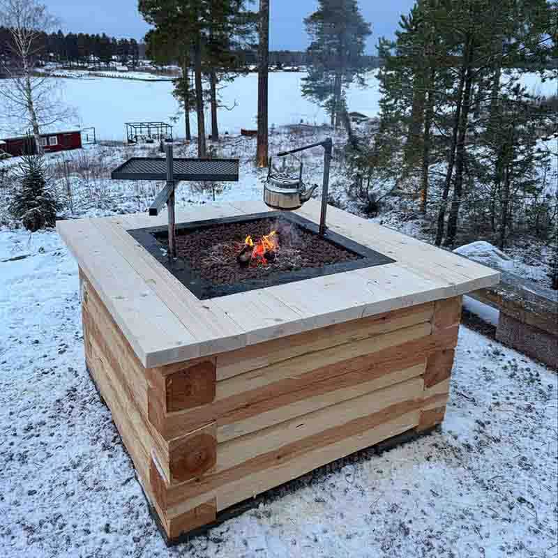 Timrad eldstad med ett bord av plankor runt omkring. I centrum en plåtinsats av corten utrustad med ett 30x40cm grillgaller och en kaffekrok där en rostfri kaffepanna hänger. En eld brinner. I bakgrunden skog och en sjö som är snö och isbelagd.