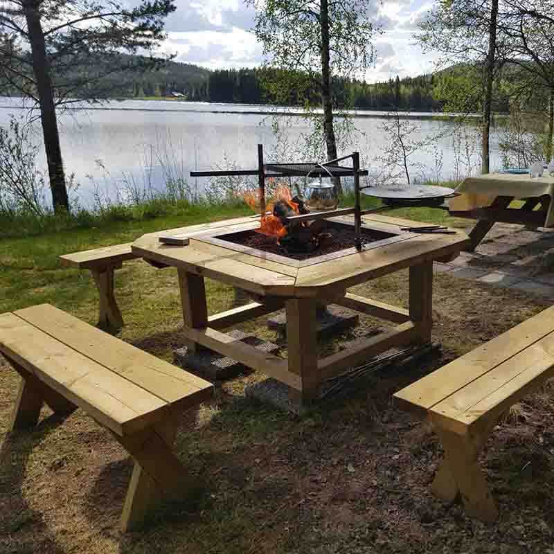 Grillplats med träbänkar runt omkring. Grillplatsen är utrustad med grillgaller, stekhällsring och kaffekrok. På kaffekroken hänger en kaffepanna och på stekhällsringen en stekhäll. I bakgrunden sommargröna björkar och en sjö.