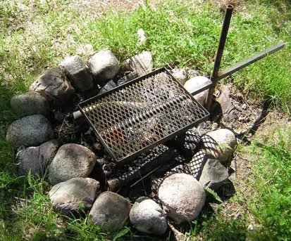 Ett rektangulärt grillgaller med handtag är monterat på ett rör som sitter fast i marken. På marken är stenar placerade i en ring för att hindra riska att elden sprider sig om man eldar under gallret.