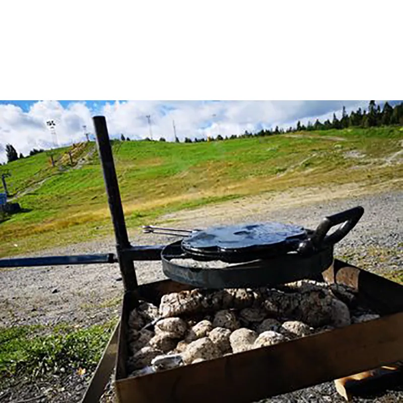 Vafflor_1_RBG Eldbox är en hopfällbar eldstad eller grill. På bilden är den uppställd på marken, det är sommar och en grön kulle i bakgrunden. I eldbox glöder det och på ett höj och sänkbart stativ ligger ett våffeljärn av gjutjärn.